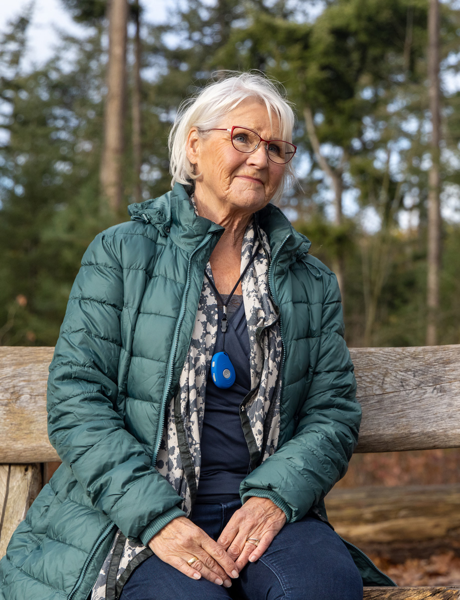 Oudere dame wandelaarster zit met een Pebbles alarm om haar nek op een bankje te genieten in het bos