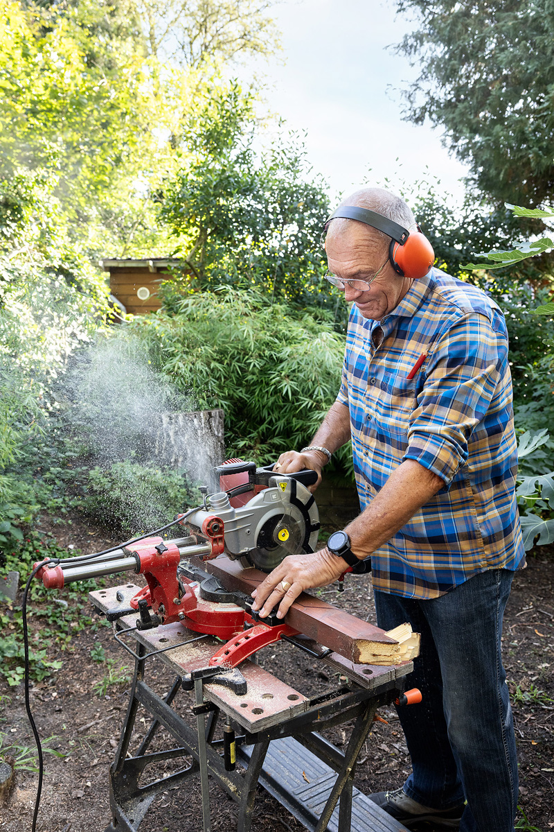 Man achter een werkbank met zaagmachine in de tuin. Aan zijn pols draagt hij een alarmhorloge.