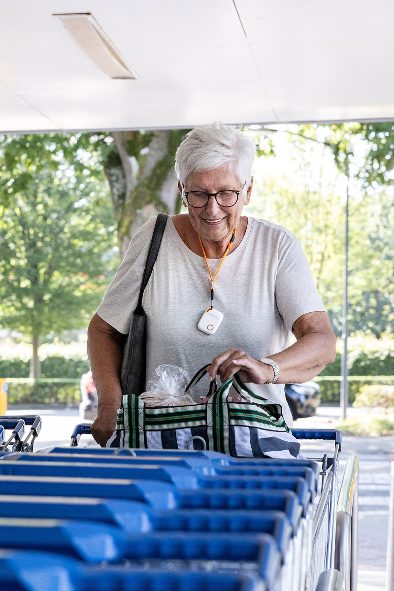 Vrouw met boodschappentas zet een winkelwagentje terug. Om haar hals draagt ze een alarmknop.