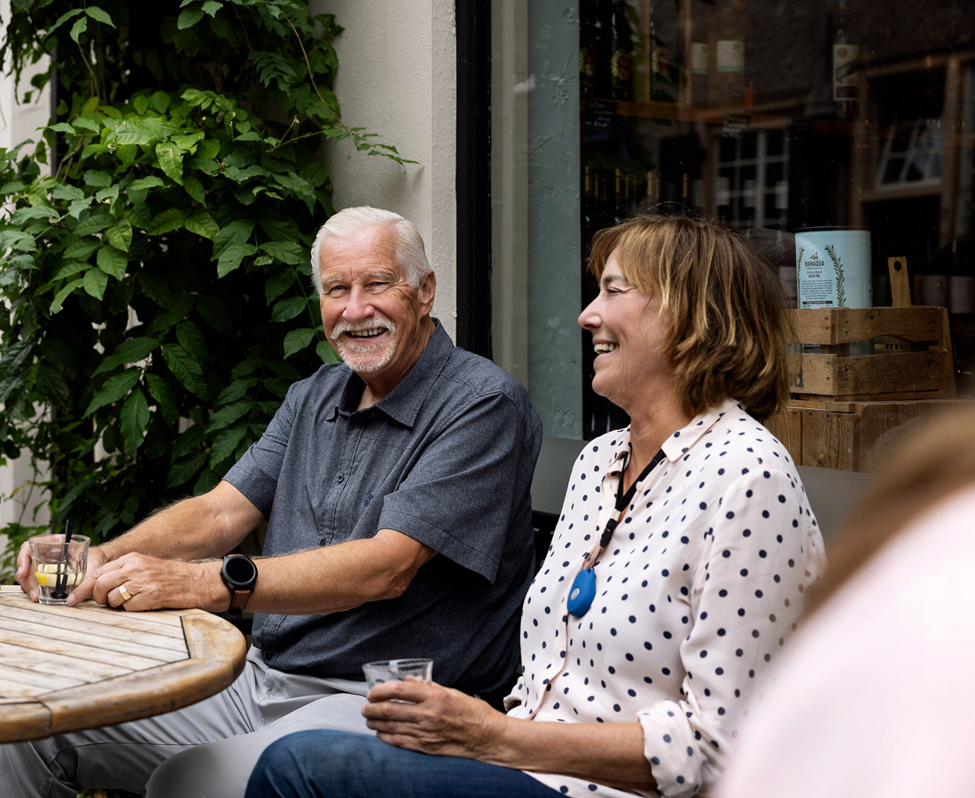 Lachend gezelschap van personen zittend aan een tuintafel. Om haar hals draagt een vrouw een alarmknop.